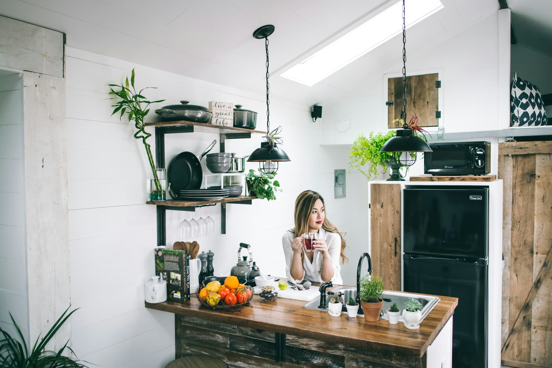 Woman seated at a kitchen island enjoying a drink, surrounded by fresh produce and potted plants