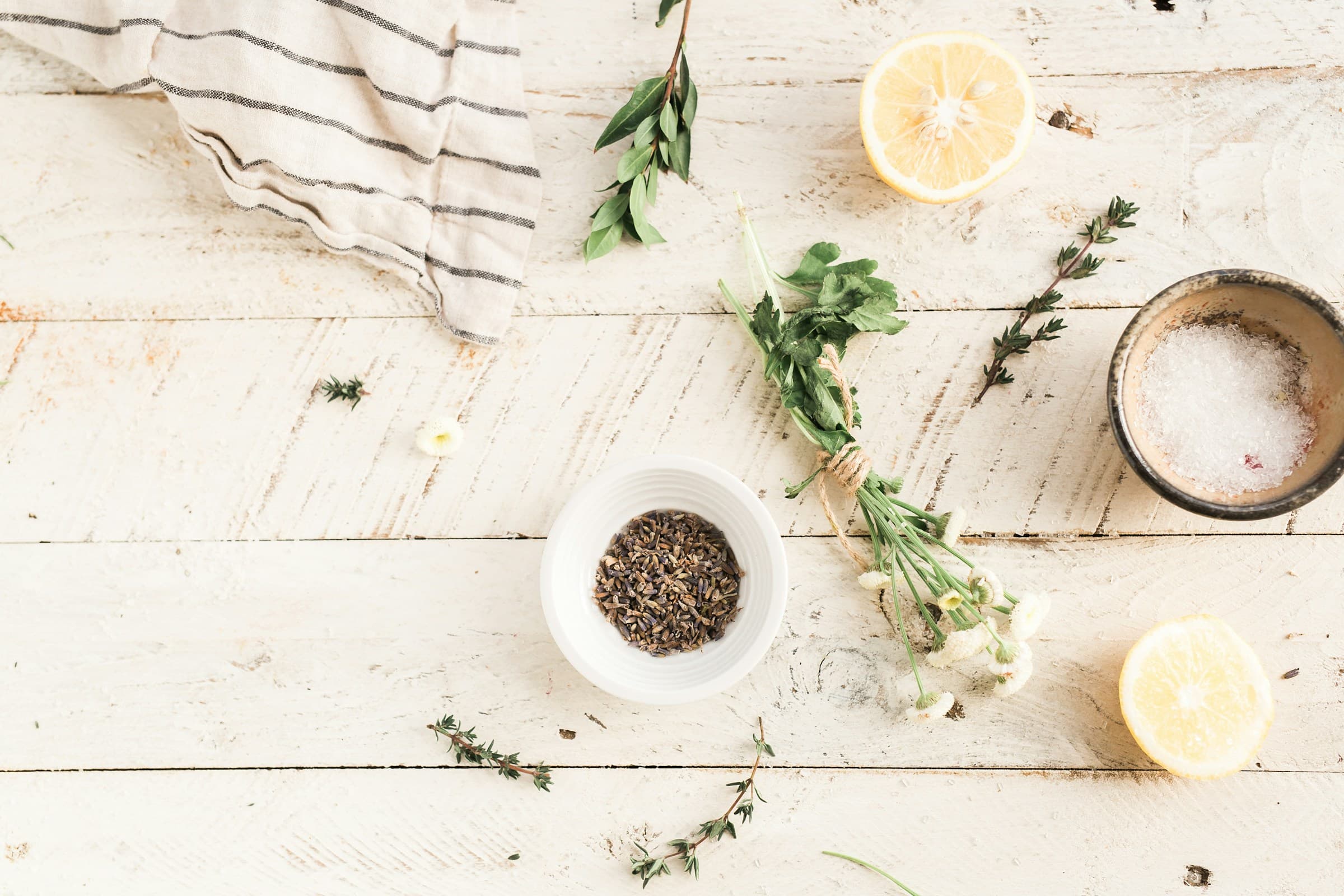 Flat lay of herbs, a lemon half, and a striped cloth on a rustic white wooden table